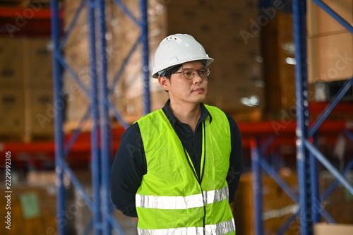 Professional Worker in Warehouse Wearing Safety Vest and Hard Hat Overseeing Inventory Stock Management Operations in Distribution Center Environment