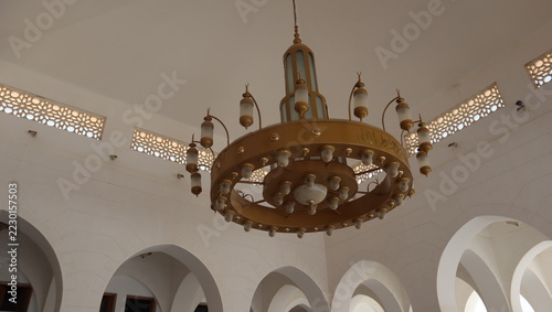 Low angle view of a large ornate golden chandelier with arabic calligraphy hanging inside a mosque with white arches.