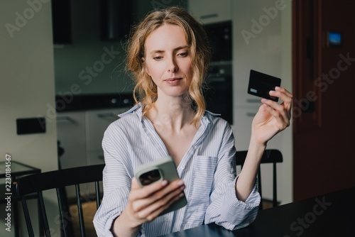 Woman holding a credit card and checking her smartphone at home, illustrating online payment fraud risks, financial scams and the danger of sharing banking information with cybercriminals