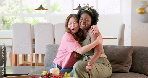 Happy black and korean lesbian couple celebrating, clapping hands and blowing candles at birthday pa