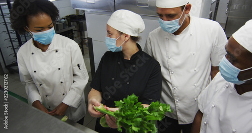 Diverse group of chefs wearing face masks looking at vegetable in restaurant kitchen