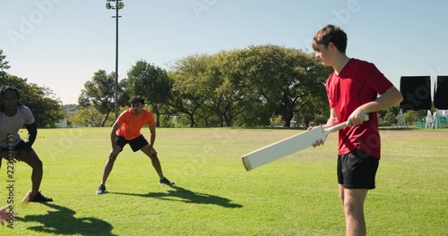 Diverse team training cricket at park, batsman bouncing red ball, swinging bat as fielders diving