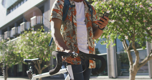 Midsection of african american man in city, wheeling bike and using smartphone in the street