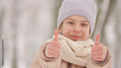 Beautiful girl in jacket and hat with a bright scarf shows thumb positively on the background of a winter city park.