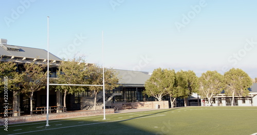 White rugby goalpost standing on green turf field, with brick wall, veranda tables and trees