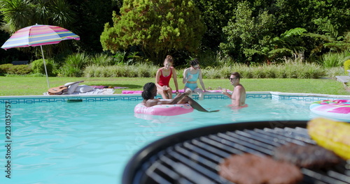 Diverse friends chatting while lounging at backyard pool beside inflatable ring and barbecue grill