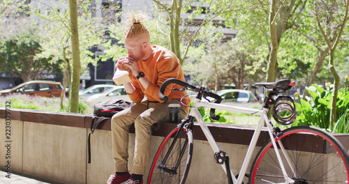 Albino african american man with dreadlocks sitting in park with bike eating sandwich
