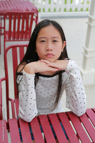 Bored Asian preteen girl sitting at red outdoor cafe table and resting her chin on her clasped hands with a serious, unhappy expression while looking at camera.