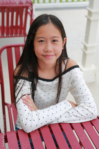 Smiling Asian preteen girl with sitting at red outdoor cafe table with her arms folded on the table and looking at camera.