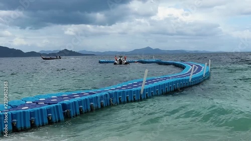 Long floating blue pier extending across turquoise seawater toward a distant tropical coastline, with small boats, green islands, and a cloudy sky