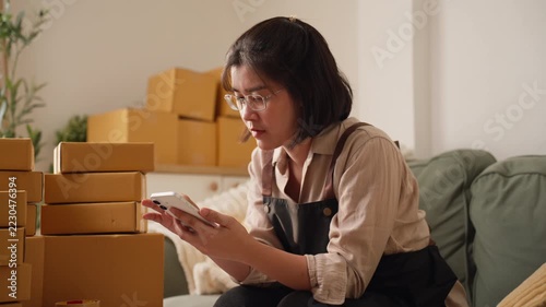Asian woman running a small online business checks her smartphone while reaching toward stacked cardboard parcels at home, illustrating ecommerce order management, inventory control, and shipping prep