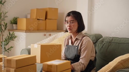 Asian woman managing a home ecommerce business reviews paperwork beside stacked cardboard shipping boxes, representing order verification, parcel preparation, inventory administration, and small busin