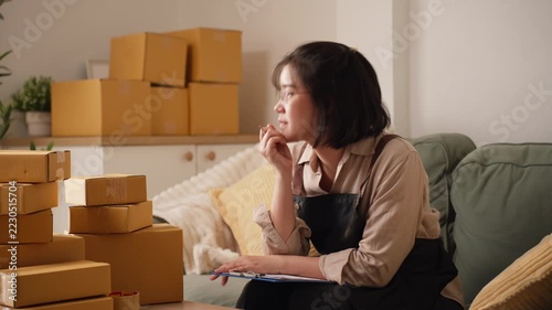 Asian female online seller wearing glasses and apron carefully checking parcel orders on clipboard file at home workstation desk.