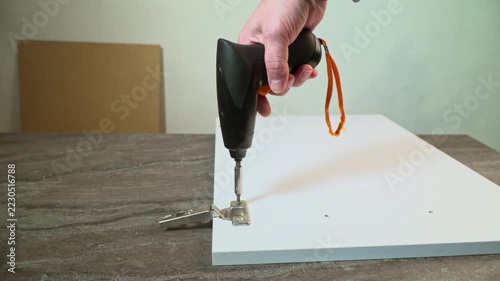 Installing metal hinge on white furniture panel with electric screwdriver. DIY furniture assembly or repair process.