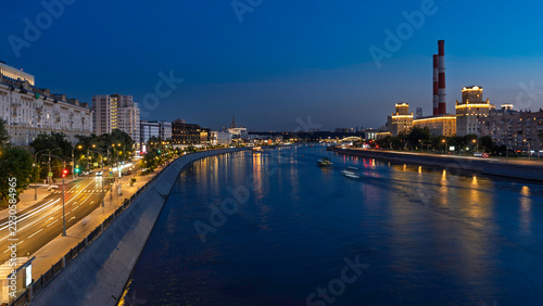 Urban river and illuminated city streets at blue hour, with traffic trails and industrial buildings along the waterfront. Cityscape and riverfront infrastructure.