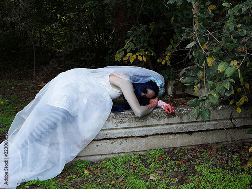 dead bride in white wedding dress lying on old stone grave in cemetery, gothic halloween horror