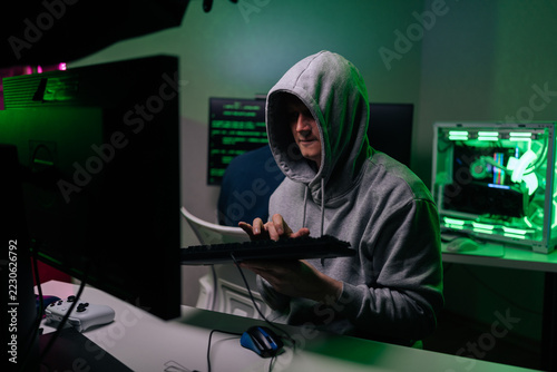 Mysterious anonymous hacker in gray hoodie at desk in dark room, typing on keyboard. Green light from monitors and PC case illuminates him. Cybersecurity and crime concept.