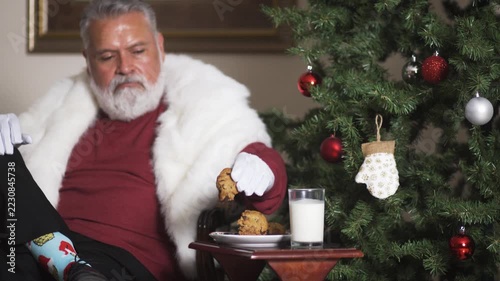 Santa dips cookie in milk and eats beside Christmas tree