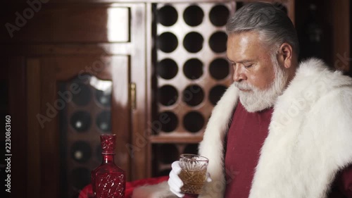 Senior, elegant Santa holds amber drink and sets glass down at home bar