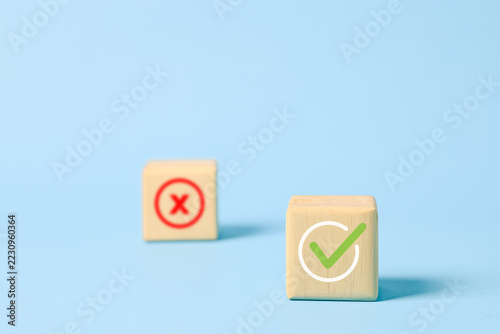 Fotografie Wooden cube with a green check mark in sharp focus and a blurred red cross symbol behind it on a pastel blue background, representing correct decisions, approval, validation and choosing success