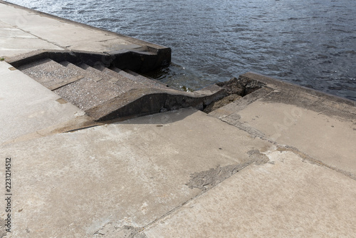 Obraz na plátně Damaged concrete riverbank shows cracked slabs and worn steps descending toward dark water