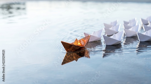 Unique paper boat sailing among white boats on tranquil water surface at sunset