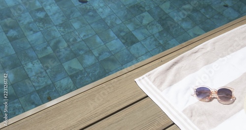 Striped towel with translucent sunglasses resting on wooden deck beside tiled pool, copy space
