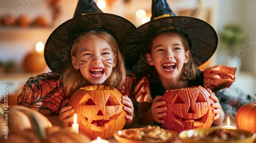 Halloween Holiday Theme. a carved pumpkin. The girl on the left wears a black witch hat with a pointed brim and a spider web pattern, and her face is painted with black catlike markings.