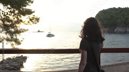 Young woman watching sailboat on calm coastal bay at sunset behind