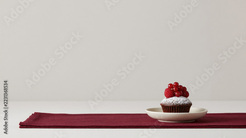 Chocolate cupcake topped with raspberries and red currants on a white plate. Minimal dessert still life with ample copy space. Bakery treat and sweet food concept. Elegant food styling.