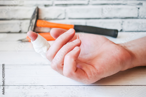 Male hand with bandaged injured finger on white wooden table with blurred hammer in background. Work injury and first aid concept for occupational safety and insurance industry.