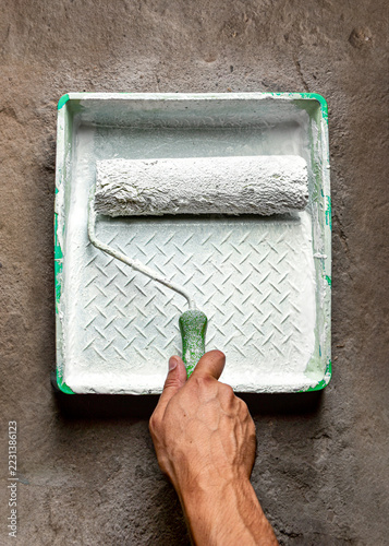 Male hand holding a green paint roller handle over a plastic tray filled with white paint on a rough concrete floor. Home renovation and construction theme in the building industry.