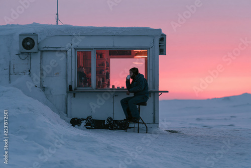 A person sits by a kiosk in the Arctic in winter, amidst the snow.