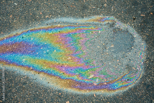 Colorful oil slick rests on rain darkened asphalt, with rainbow film outlining shallow water and scattered grit. Vivid iridescent pattern offers abstract view of urban road surface pollution.