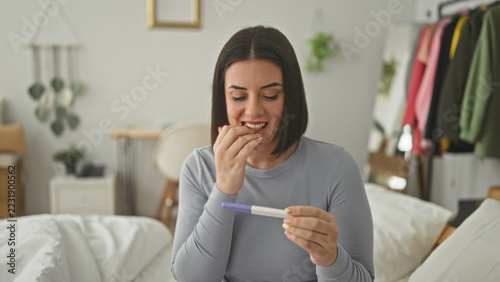 Obraz na plátně Woman young with short hair holding a pregnancy test, biting nails and fingers crossed while seated on a bed in a building; hope anticipation