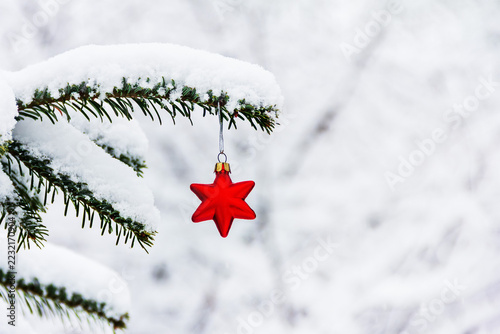 A bright red Christmas tree ornament in the shape of a star hangs from a snow‑covered spruce branch, giving the winter landscape a festive look.