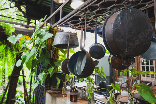 Obraz na plátně Hanging vintage metal pots and pans dangle from a metal rack in a lush, plant-filled rustic outdoor kitchen space