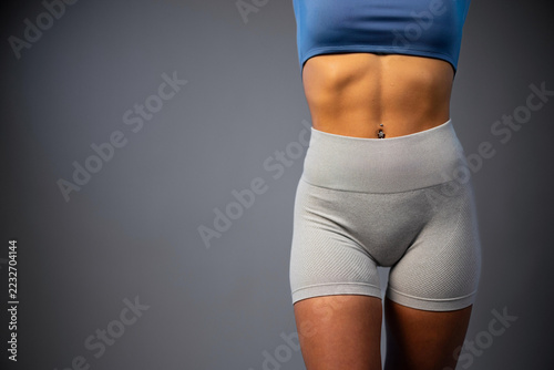 Fit woman in blue sports bra and gray leggings showing defined abs on gray background