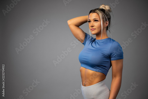 Fit young woman in blue crop top and gray leggings posing confidently against gray background