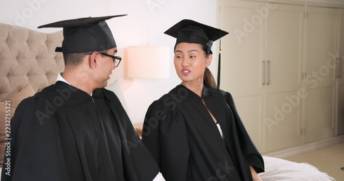 Young couple in graduation gowns adjusting caps at home, preparing for ceremony