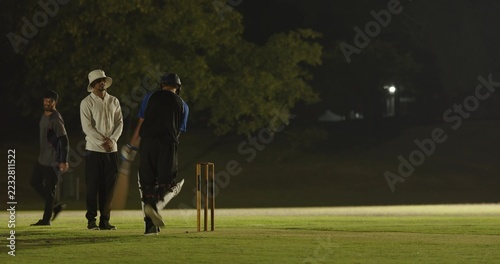 Bowler returning to crease on floodlit field prompts batsman practicing swinging bat, bumping fists