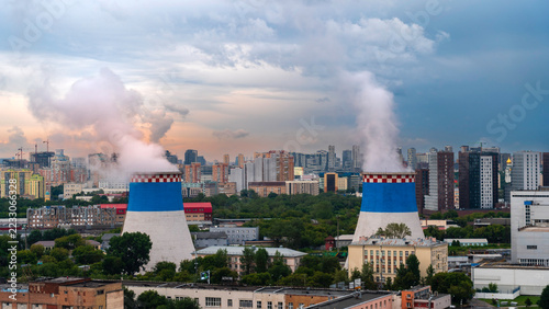Power plant cooling towers releasing steam above dense urban city buildings. Electricity generation and energy infrastructure. Industrial pollution and environmental impact.