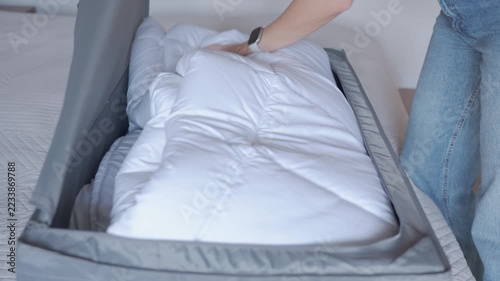 Woman packing white duvet into gray storage bag on bed. Hands arranging bedding inside and closing bag with zipper