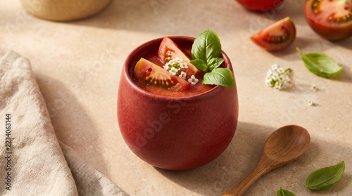 Red apple shaped bowl filled with creamy tomato soup and basil garnish on a warm natural table with fresh tomatoes wooden spoon and rustic linen for healthy seasonal food styling