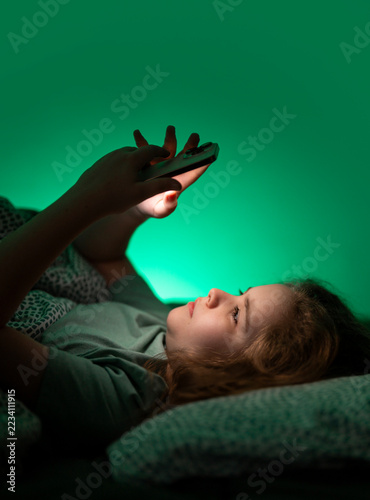 Child playing on phone in bed at night. Kid resting in bed with phone at night. Teenager watching media on phone in bed at night. Child focused on phone while in bed at night.