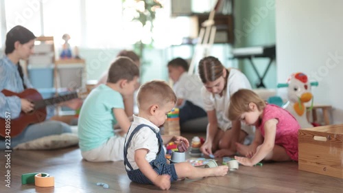 Toys are scattered on floor, children are playing with cubes and cars, teacher is playing guitar. Girl babysitter accompanies children, twang guitar during childrens play with toys activities. 