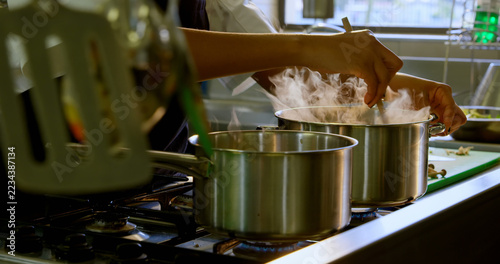 A person is cooking, stirring a pot on a stove with steam rising from the pans, with copy space
