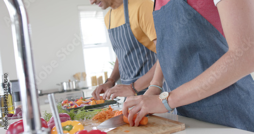 Happy diverse couple preparing fresh vegetables and wearing aprons in kitchen, slow motion