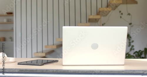 Silver laptop sitting on wooden desk by staircase, black tablet with headphones near trailing plant