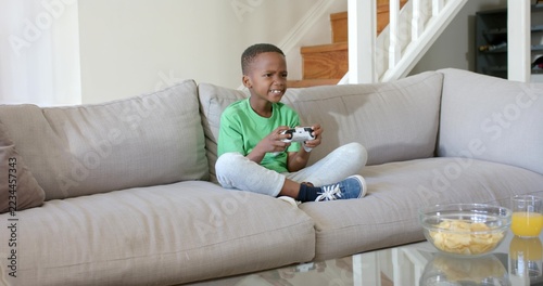 Playing boy wearing green tee holding wireless controller on sofa at home, with chips, orange juice